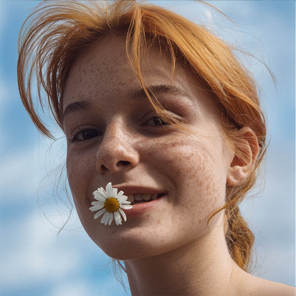Smiling red-haired woman with freckled skin holding a daisy in her mouth representing natural care with grass-fed tallow balm